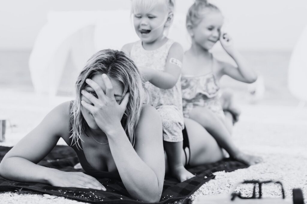 a mum looks overwhelmed as she lies on the floor with two young children sitting on her back